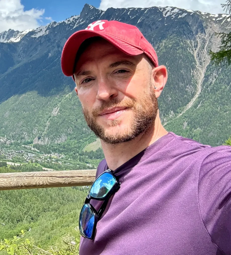 Dave Bell taking a selfie at a mountain viewpoint, wearing a red cap and purple shirt, with alpine peaks and a green valley behind him.
